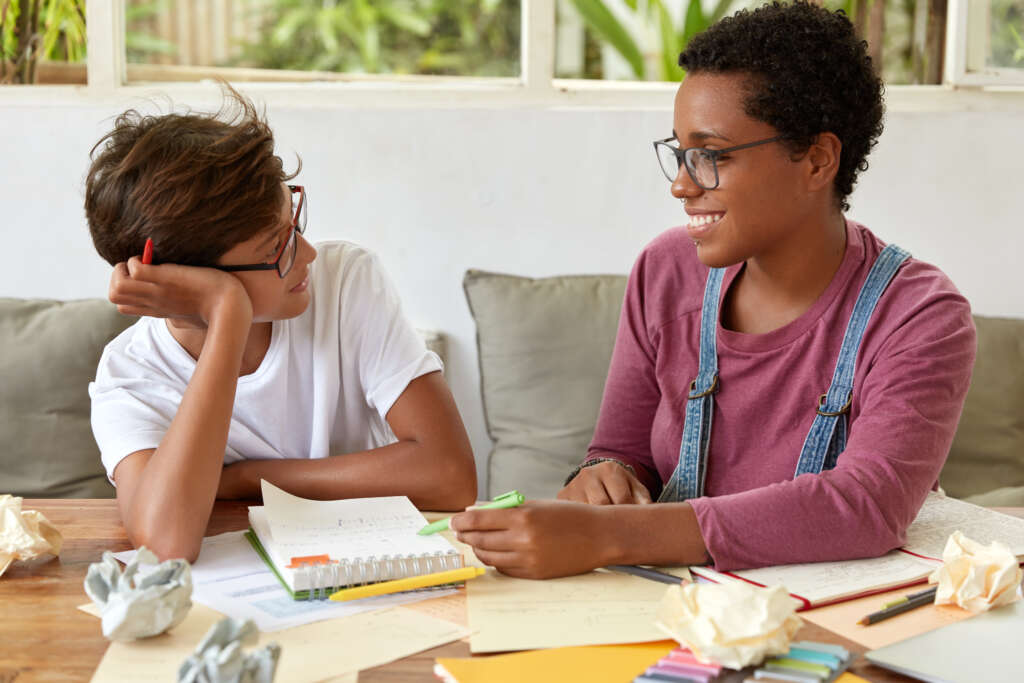 horizontal shot of mixed race women have conversation during learning process, sit on couch, look happily, wear spectacles, surrounded with papers, have positive expressions, discuss ideas for project